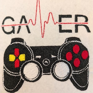 Puede incluir: Un diseño bordado sobre una superficie de tela blanca que muestra la palabra GAMER en letras mayúsculas negras con una línea de latido roja en el centro. Debajo hay un mando de videojuego negro con botones rojos y amarillos.