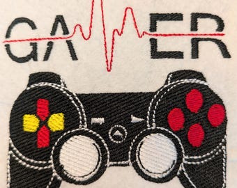 Peut inclure: Un motif brodé sur un tissu blanc représentant le mot GAMER en lettres capitales noires avec une ligne de rythme cardiaque rouge au centre. En dessous se trouve une manette de jeu vidéo noire avec des boutons rouges et jaunes.