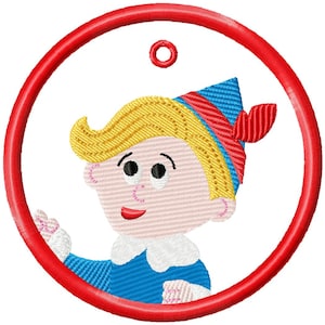 Op de afbeelding: Geborduurd ontwerp van een stripfiguur met blond haar, een blauwe hoed en een rode strik, geplaatst in een rood cirkelvormig frame. Het personage draagt een blauw shirt met een witte kraag en glimlacht. Het ontwerp staat op een witte achtergrond.