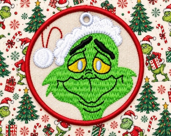 May include: A circular embroidered ornament featuring a green character with yellow eyes and black eyebrows, wearing a white Santa hat. The design is stitched onto a light-coloured fabric background with a red border and a white hanging loop.