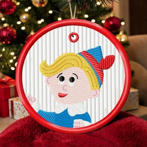 Op de afbeelding: Een ronde kerstversiering met een rode rand en een cartoon elf ontwerp. De elf heeft blond haar, een blauwe hoed met rode en blauwe strepen en een blauw shirt. De achtergrond is wit met verticale lijnen.