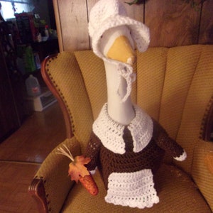 May include: A crocheted goose wearing a white crocheted hat and a brown crocheted dress sitting in a yellow armchair.