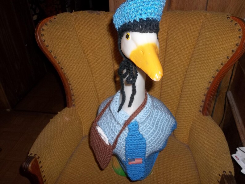 May include: A white goose figurine wearing a blue crocheted hat, a blue crocheted tie, and a brown crocheted vest. The goose is sitting on a brown armchair.