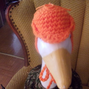 Puede incluir: Un gorro de punto naranja sobre una figura de ganso de madera. El ganso tiene la cabeza blanca y un pico largo de madera.