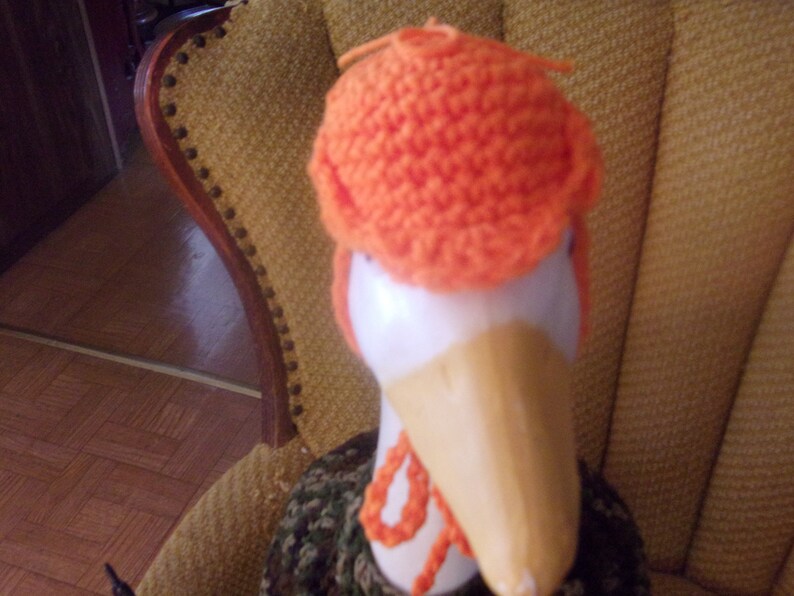 Puede incluir: Un gorro de punto naranja sobre una figura de ganso de madera. El ganso tiene la cabeza blanca y un pico largo de madera.