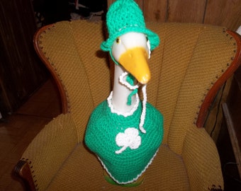 Puede incluir: Un ganso con un sombrero y capa de ganchillo verdes, adornado con un trébol blanco. El ganso tiene un pico amarillo y está sentado en un sillón vintage.