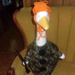 Puede incluir: Un ganso blanco con un su&eacute;ter de camuflaje y un gorro de crochet naranja. El ganso est&aacute; sentado en una silla amarilla.