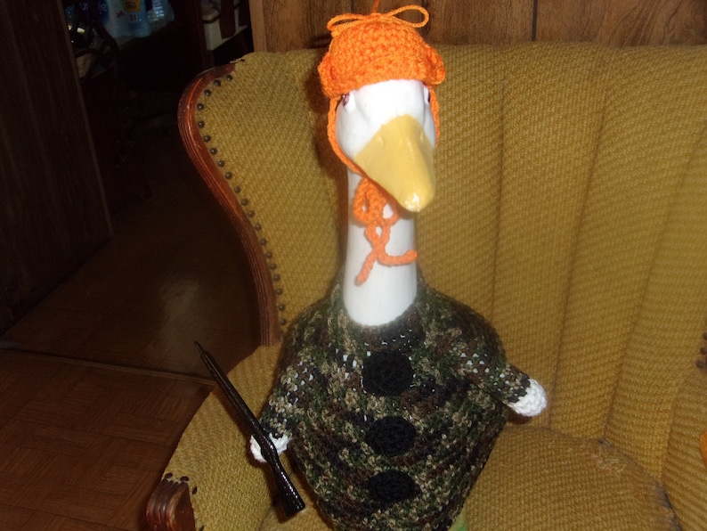 Puede incluir: Un ganso blanco con un su&eacute;ter de camuflaje y un gorro de crochet naranja. El ganso est&aacute; sentado en una silla amarilla.