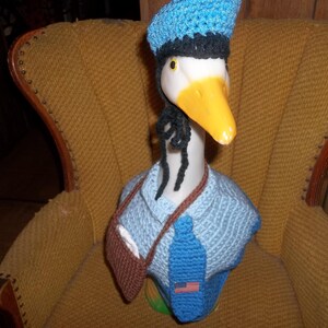 May include: A white goose figurine wearing a blue crocheted hat, a blue crocheted tie, and a brown crocheted vest. The goose is sitting on a brown armchair.