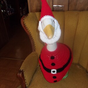 May include: A goose wearing a handmade Santa Claus outfit, including a red hat, beard, and jacket. The goose is sitting in a vintage yellow armchair. The outfit has black accents and a white belt.