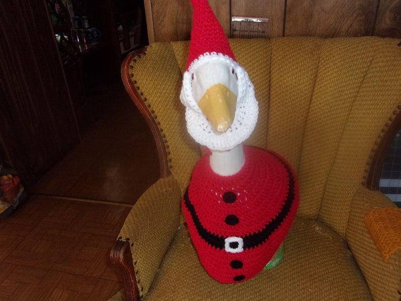 May include: A goose wearing a handmade Santa Claus outfit, including a red hat, beard, and jacket. The goose is sitting in a vintage yellow armchair. The outfit has black accents and a white belt.