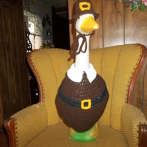 May include: A white goose wearing a brown crocheted pilgrim hat and a brown crocheted vest with a yellow buckle. The goose is sitting on a yellow armchair.