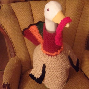 May include: A white goose wearing a crocheted turkey costume. The costume is brown, orange, red, and green. The goose is sitting in a brown armchair.