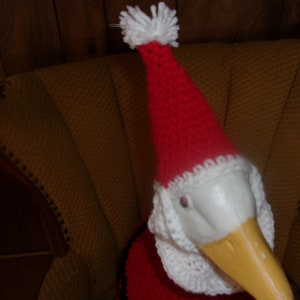 May include: A goose figurine wearing a red and white Santa Claus hat and outfit. The goose has a yellow beak and is sitting in a yellow armchair. The hat has a white pom-pom on top.