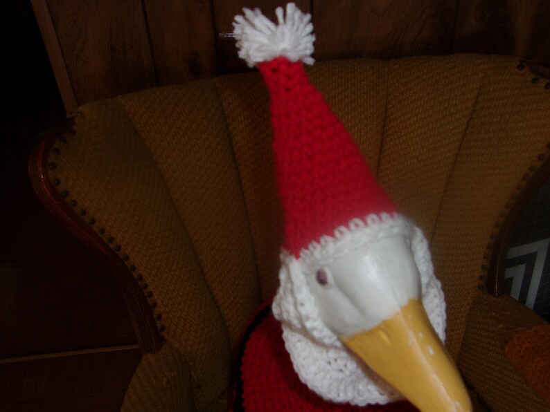 May include: A goose figurine wearing a red and white Santa Claus hat and outfit. The goose has a yellow beak and is sitting in a yellow armchair. The hat has a white pom-pom on top.