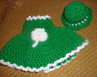 Puede incluir: Un vestido y sombrero de ganchillo verdes hechos a mano, ambos con bordes blancos. El vestido presenta un trébol blanco. El conjunto es probablemente para un bebé o niño pequeño, ideal para el Día de San Patricio.