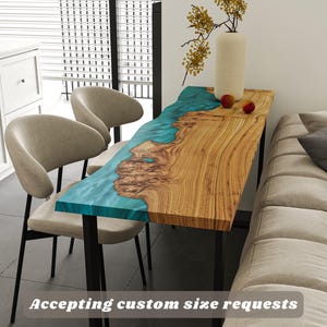 May include: A wooden console table with a turquoise resin river design. The table has a natural wood grain finish and black metal legs. Two beige chairs are in front of the table. The text "Accepting custom size requests" is at the bottom.