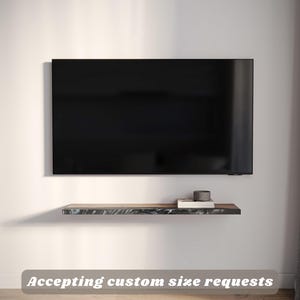 May include: A black television is mounted on a white wall above a floating shelf. The shelf has a dark, textured surface with a book and a small, dark grey cylindrical object. The text "Accepting custom size requests" is displayed.
