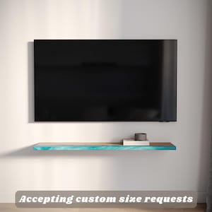 May include: A wall-mounted wooden shelf with a turquoise and brown design is positioned below a black television. The shelf holds a small stack of books and a dark grey cylindrical object. The text "Accepting custom size requests" is displayed.