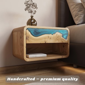 Könnte beinhalten: Ein schwebender Nachttisch aus Holz mit einer Schublade und einem offenen Regal. Die Schubladenfront hat eine blaue Harzeinlage. Oben steht eine kleine Vase mit Trockenblumen. Der Text "Handcrafted - premium quality" befindet sich unten.