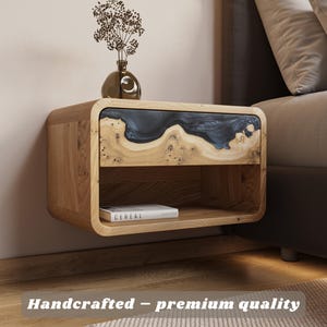 May include: A floating wooden nightstand with a drawer featuring a dark blue resin inlay. The nightstand has a lower shelf with a book titled "Cereal". A small vase with dried flowers sits on top. The text "Handcrafted - premium quality" is at the bottom.