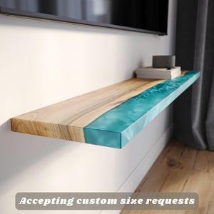 May include: A wooden floating shelf with a turquoise resin river design. The shelf is fixed to a white wall. The wood has a natural grain. The shelf holds books and a small black box. Text on the image says "Accepting custom size requests."