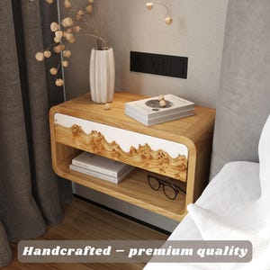 May include: A wooden nightstand with a curved design, featuring a white accent and a lower shelf. A vase with dried flowers, a book, and eyeglasses are on the nightstand. The text "Handcrafted - premium quality" is visible.