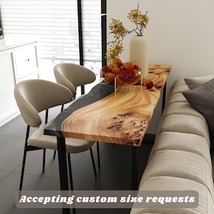 May include: A live edge dining table with a black resin inlay and black metal legs. The table is made of light brown wood and has a natural wood grain pattern. The text "Accepting custom size requests" is visible.