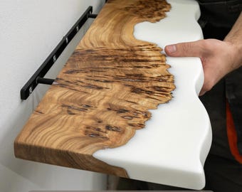 May include: A rectangular wall shelf featuring a natural wood slab with a prominent grain pattern on one side and a smooth, opaque white resin section on the other. The shelf is attached to a white wall with a black metal bracket system.