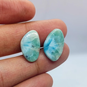 May include: Two polished larimar gemstones, each with a unique pattern of blue and white swirls on a light teal background. The stones are held between fingers, showcasing their natural beauty and potential for jewelry making.