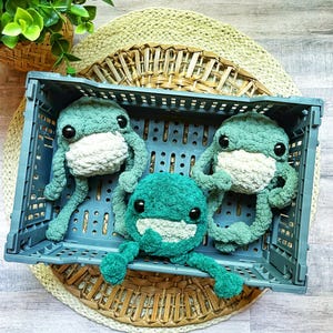 May include: Three crocheted green frog plushies with white faces and eyes, sitting inside a gray plastic crate. The crate is on top of a woven straw mat.