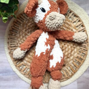 May include: A crocheted plush toy cow with brown and white fur. The cow has a brown nose and black eyes. The toy is lying on a woven basket.