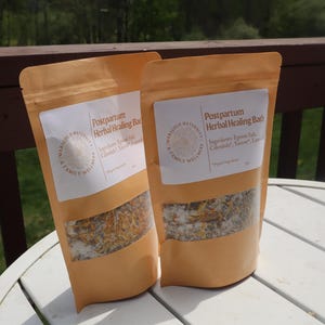 Può includere: Due sacchetti beige di bagno curativo a base di erbe post-partum sono esposti su un tavolo bianco. Ogni sacchetto ha un'etichetta bianca con testo e una finestra trasparente che mostra il contenuto, tra cui sale di Epsom e calendula. Lo sfondo comprende un terrazzo in legno e vegetazione.