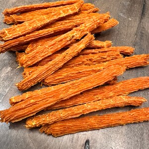 K&ouml;nnte beinhalten: Ein Haufen orangefarbener, d&uuml;nner, langer und leicht gekr&auml;uselter Hunde-Leckerlis. Die Leckerlis sind auf einer Holzoberfl&auml;che zu einem Haufen gestapelt.