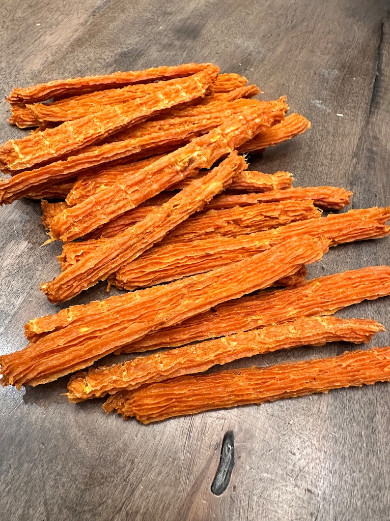K&ouml;nnte beinhalten: Ein Haufen orangefarbener, d&uuml;nner, langer und leicht gekr&auml;uselter Hunde-Leckerlis. Die Leckerlis sind auf einer Holzoberfl&auml;che zu einem Haufen gestapelt.
