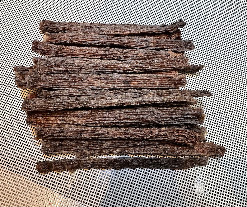 K&ouml;nnte beinhalten: Nahaufnahme eines Stapels d&uuml;nner, dunkelbrauner Rindfleisch-Jerky-Streifen auf einer wei&szlig;en Netzfl&auml;che.