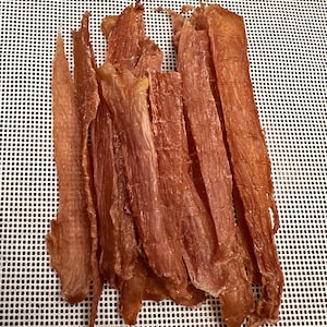 May include: A close-up shot of several pieces of dried meat, likely jerky, arranged on a white, perforated surface. The meat strips are a reddish-brown color, with visible textures and fibers. The lighting highlights the details of the dried meat.