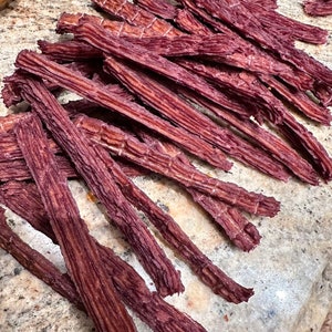 K&ouml;nnte beinhalten: Ein Haufen d&uuml;nner, langer, dunkelroter Rindfleisch-Jerky-Streifen. Der Jerky ist in einem zuf&auml;lligen Muster auf einer hellbraunen Oberfl&auml;che angeordnet.