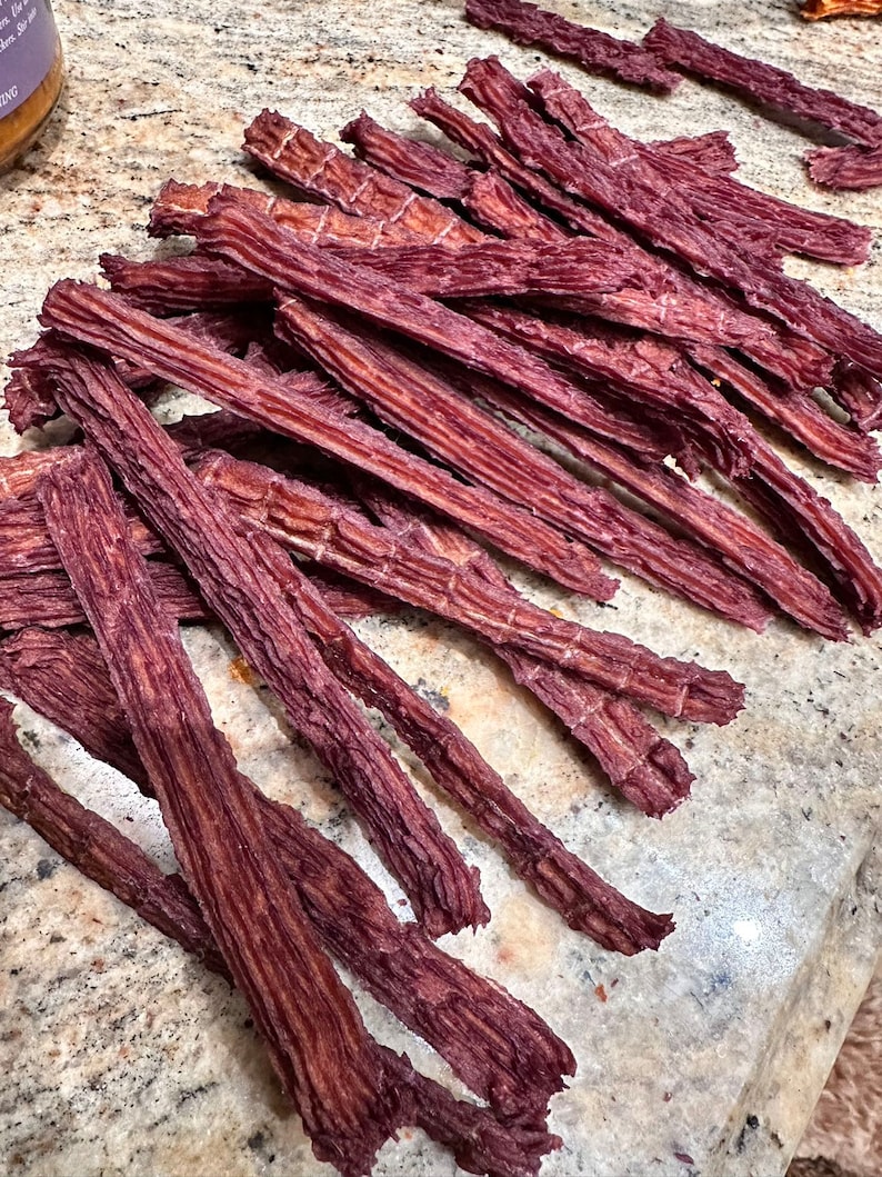 K&ouml;nnte beinhalten: Ein Haufen d&uuml;nner, langer, dunkelroter Rindfleisch-Jerky-Streifen. Der Jerky ist in einem zuf&auml;lligen Muster auf einer hellbraunen Oberfl&auml;che angeordnet.