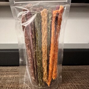 K&ouml;nnte beinhalten: Ein transparenter Plastikbeutel gef&uuml;llt mit verschiedenen bunten, langen, d&uuml;nnen, getrockneten Fleischleckerlis. Die Leckerlis sind vertikal im Beutel angeordnet, wobei die Enden der Leckerlis oben sichtbar sind.