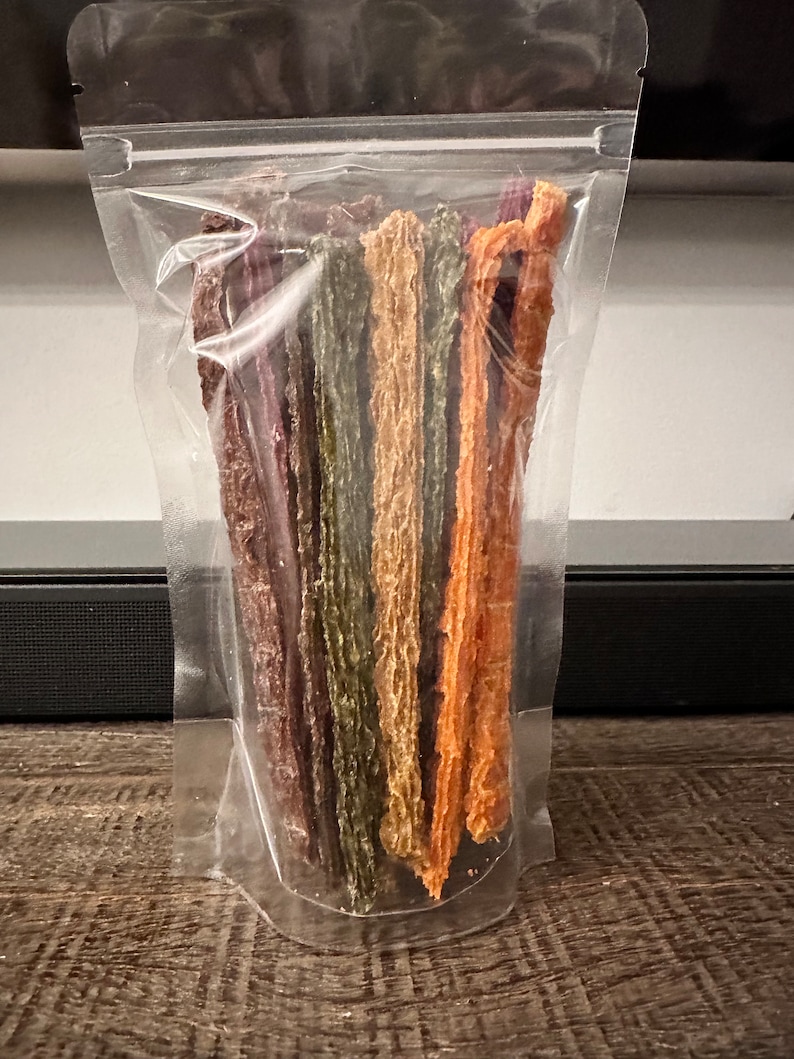 K&ouml;nnte beinhalten: Ein transparenter Plastikbeutel gef&uuml;llt mit verschiedenen bunten, langen, d&uuml;nnen, getrockneten Fleischleckerlis. Die Leckerlis sind vertikal im Beutel angeordnet, wobei die Enden der Leckerlis oben sichtbar sind.