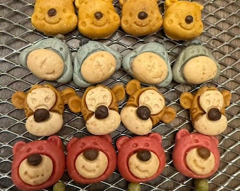 May include: A variety of Disney-themed cookies arranged on a wire rack. The cookies feature characters like Winnie the Pooh, Piglet, Tigger, Lotso, and the Little Green Men, in various colors including pink, yellow, and green.