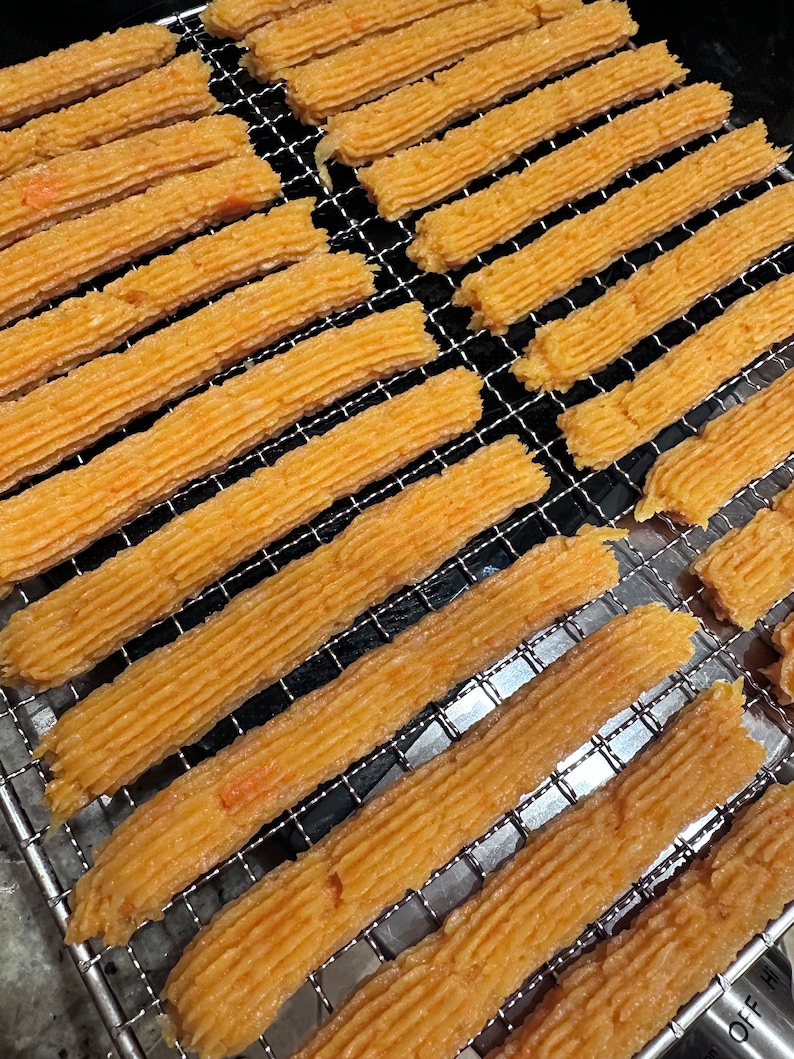 K&ouml;nnte beinhalten: Nahaufnahme eines Drahtgestells mit langen, d&uuml;nnen, orangefarbenen Streifen aus Essen. Die Streifen sind in Reihen angeordnet und leicht gekr&uuml;mmt.