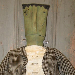 Puede incluir: Una muñeca de tela con cabeza rectangular verde, detalles faciales cosidos y parte superior plana. Viste una chaqueta de cuadros negros y blancos sobre un chaleco color crema sujeto con imperdibles metálicos y pantalones negros.