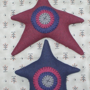 May include: Two felt star ornaments with a concentric circle design. The larger star is burgundy with a navy blue circle and the smaller star is navy blue with a burgundy circle. Both stars have a gray center circle.