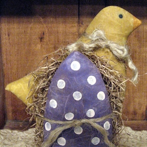May include: A purple egg with white polka dots is nestled in straw and surrounded by a yellow chick with a brown twine bow.