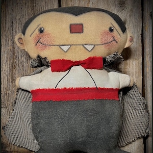 Könnte beinhalten: Handgemachte Vampir-Plüschpuppe mit beigem Gesicht, schwarzen Haaren und roter Fliege. Die Puppe trägt ein weißes Hemd, eine graue Hose und einen schwarz-weiß gemusterten Umhang. Der Vampir hat ein gesticktes Lächeln und zwei weiße Fangzähne.