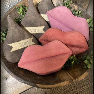 Puede incluir: Un cuenco de madera contiene objetos decorativos: tres adornos en forma de Kiss de Hershey's de color marrón oscuro con pancartas "KISS", y tres labios de felpa en tonos de rosa y rojo. La vegetación acentúa la disposición. El texto "Chestnut Junction" está en la parte inferior.