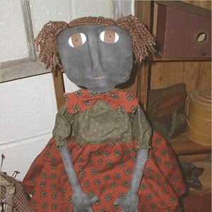Może przedstawiać: Ręcznie robiona lalka ze szmatki z brązowymi włosami z wełny, szarą twarzą i brązowymi oczami. Lalka ma na sobie czerwoną i zieloną sukienkę w kwiaty i siedzi na drewnianym krześle. Lalka jest oznaczona "Chestnut Junction".
