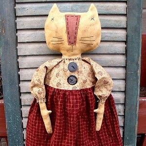 May include: A handmade rag doll cat with a tan face, red nose, and black eyes. The doll is wearing a brown and gold dress with a red and white checked skirt. The doll is standing on a wooden surface.